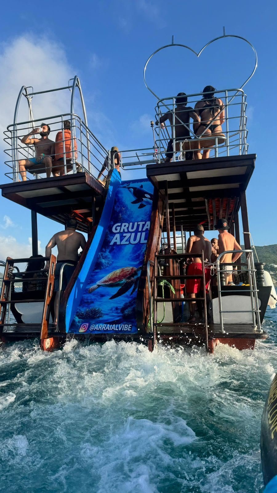Passageiros na popa da embarcação Gruta Azul com moldura em formato de coração e escorregador ao fundo